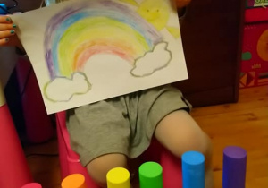 dziewczynka pokazuje pokolorowany obrazek z tęczą, obok kolorowe klocki